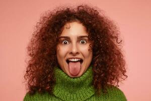 Young lady, funny woman with ginger curly hair. Wearing green turtleneck sweater and showing her tongue. Watching at the camera with big eyes, isolated, closeup over pastel pink background photo