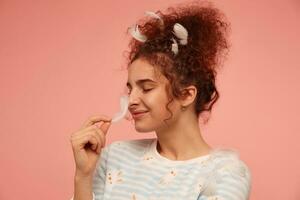 Young lady, lovely woman with ginger curly hair. Wearing striped sweater with bunnies and covered with feathers, touching her nose with feather. Stand isolated, closeup over pastel pink background photo