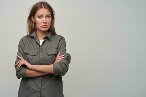 Portrait of attractive, adult girl with brown long hair. Wearing grey shirt and holding arms crossed on a chest. Watching sad at the camera, copy space at the right, isolated over grey background photo