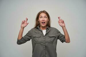 Teenage girl, happy looking woman with brown long hair. Wearing grey shirt and keep her fingers crossed, making a wish, asking. Watching up at copy space, isolated over grey background photo