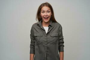 Teenage girl, happy looking woman with brown long hair. Wearing grey shirt and surprised, shocked. Happy to see you. Watching at the camera with big eyes, isolated over grey background photo