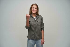 Teenage girl, happy looking woman with brown long hair. Wearing grey shirt and jeans. Emotional concept. Keep fist lifted and squints. Watching at the camera isolated over grey background photo