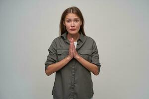Teenage girl, unhappy woman with brown long hair. Wearing grey shirt and keep her palms together, asks for something. Watching at the camera isolated over grey background photo