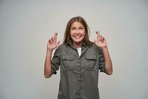 Portrait of attractive, adult girl with brown long hair. Wearing grey shirt and keep her fingers crossed. Within temptation. Watching up at copy space, isolated over grey background photo