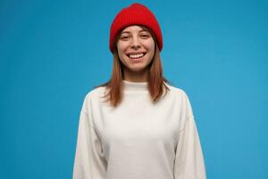 Teenage girl, happy looking woman with brunette hair. Wearing white sweater and red hat. Have a big smile. People concept. Watching at the camera, isolated over blue background photo