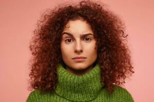Young lady, pretty woman with ginger curly hair. Serious looking. Wearing green turtleneck sweater and watching at the camera with eyebrow lifted, isolated, closeup over pastel pink background photo
