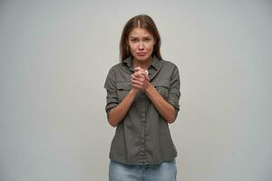 Young lady, cute woman with brown long hair. Wearing grey shirt and jeans. Keep her hands together and begs you. Watching upset at the camera isolated over grey background photo