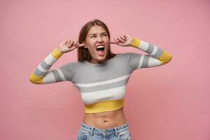 Teenage girl, angry looking european woman with brown long hair. Wearing striped blouse and jeans. Close her ears with fingers, screams. Watching up at copy space, isolated over pastel pink background photo