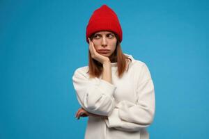 Sad looking woman, beautiful girl with brunette hair. Wearing white sweater and red hat. Put her head on a hand. Feels bored. Watching to the left at copy space, isolated over blue background photo