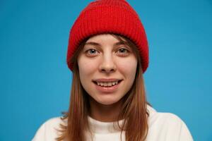 frio mirando mujer, contento niña con morena cabello. vistiendo blanco suéter y rojo sombrero. personas y emocional concepto. acecho a el cámara con un sonrisa, de cerca, aislado terminado azul antecedentes foto