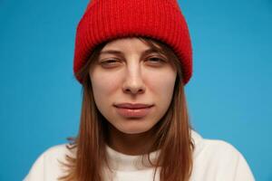 Teenage girl, suspiciously looking woman with brunette hair. Wearing white sweater and red hat. People and emotional concept. Watching at the camera, closeup, isolated over blue background photo