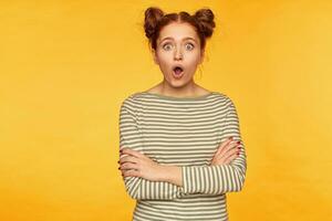 Teenage girl, shocked looking red hair woman with two buns. Wearing striped sweater and watching at the camera with hands crossed on a chest and surprised face. Stand isolated over yellow background photo