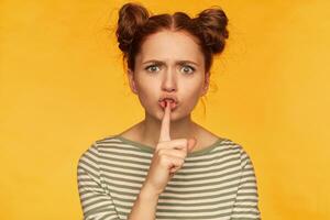 Portrait of attractive, adult girl with two buns and big eyes. Wearing striped blouse and showing silence sign, looking angry, unsatisfied. Watching at the camera isolated over yellow background photo