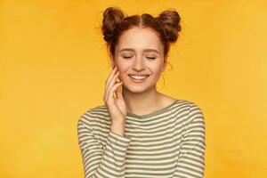 Portrait of attractive, charming red hair girl with two buns. Wearing striped sweater and smile with closed eyes, touching her cheek with fingertips. Stand isolated over yellow background photo