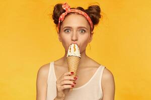 Portrait of attractive, lovely girl with two buns. Wearing white tank top and red doted hairband. Eat ice cream, watching at you with big eyes. Watching at the camera isolated over yellow background photo