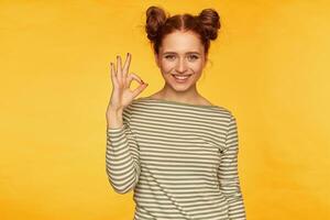 Teenage girl, happy, successful looking red hair woman with two buns. Wearing striped sweater and showing okay sign, smile. Watching at the camera isolated over yellow background photo