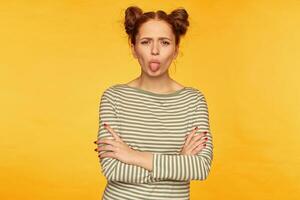 Portrait of attractive, red hair girl with two buns. Wearing striped sweater and showing a tongue to the camera with hands crossed on a chest. Stand isolated over yellow background photo