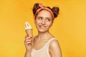 Young lady, pretty ginger woman with two buns. Wearing white tank top and red doted hairband. Holding ice cream and biting a lip. Watching to the right at copy space, isolated over yellow background photo