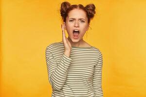 Teenage girl, unsatisfied looking red hair woman with two buns. Demonstrating her disappointed feelings. Wearing striped sweater and shout loud at the camera isolated over yellow background photo