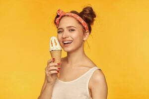 Teenage girl, happy looking red hair woman with two buns. Wearing white tank top and red doted hairband. Holding ice cream next to her face. Watching at the camera isolated over yellow background photo