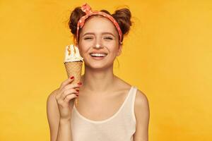 Teenage girl, red hair woman with two buns. Wearing white tank top and red doted hairband. Holding ice cream and get her nose dirty in it. Watching at the camera isolated over yellow background photo