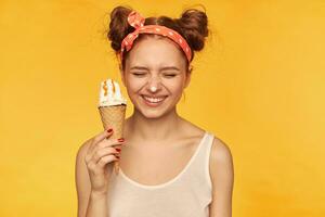 Young lady, pretty ginger woman with two buns. Wearing white tank top and red doted hairband. Holding ice cream and have it on her nose, smiling. Stand isolated over yellow background photo