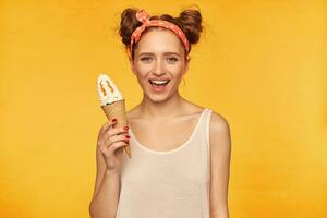 Teenage girl, happy looking red hair woman with two buns. Wearing white tank top and red doted hairband. Holding ice cream and have a big smile. Watching at the camera isolated over yellow background photo