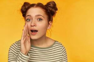 Teenage girl, happy looking red hair woman with two buns. Wearing striped blouse and holding hand next to her mouth, whisper to you. Watching at the camera isolated over yellow background photo