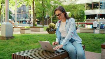 despreocupado negocio mujer se sienta al aire libre con café y utilizando computadora portátil, remoto trabajo video