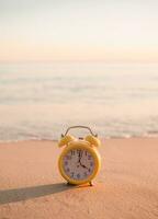 amarillo alarma reloj en el playa en el puesta de sol tiempo. el concepto acerca de hora a verano, viajar, vacaciones y relajación foto