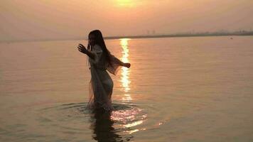 hermosa niña con largo pelo en un playa sayo bailando en pie en el agua en contra un puesta de sol antecedentes. lento movimiento. video