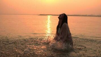 Attractive girl with long hair makes a spray of hands standing in the water against the background of the sun having a good mood. slow motion. HD video