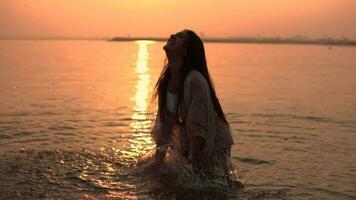 mooi meisje met lang haar- maakt een verstuiven van handen staand in de water tegen de achtergrond van de zon. langzaam beweging video