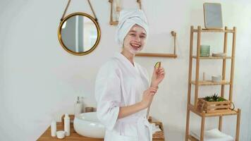 peau se soucier, après bain, produits de beauté à maison, caucasien femme, anti vieillissement. caucasien femme dans une peignoir et une cosmétique masque sur sa visage tromper autour appliquant concombres à sa yeux video