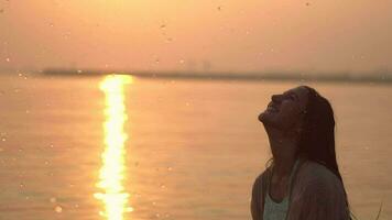 hermosa contento niña con largo pelo haciendo salpicaduras manos en pie en el río en contra el Dom durante puesta de sol teniendo un bueno humor. lento movimiento. de cerca. hd. contento día video