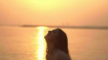 Attractive young girl with long hair makes a spray of hands standing in the water against the background of the sun. slow motion. video