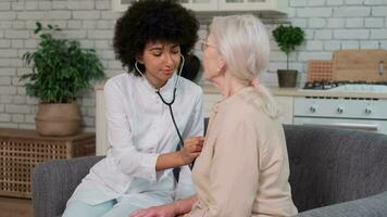 Afro american woman doctor listens to breath of senior woman using stethoscope while sitting on sofa at home. Family Doctor, Patient Support, Help at Home, Caring for the Sick. video