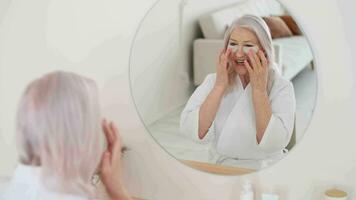 Elderly woman in robe uses eye patches while sitting in front of a mirror video