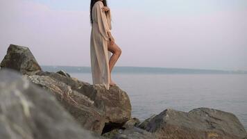 Sexy girl model with long hair and in a beach tunic is standing on stones against the background of the sea and posing. slow motion. 4K video