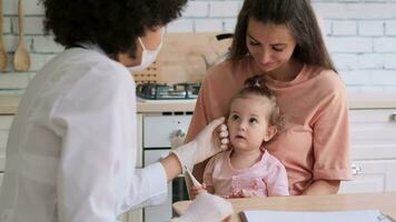 Afro american woman doctor in mask in a dressing gown supports a sick little girl while sitting at the table at home. Family Doctor, Patient Support, Help at Home, Caring for the Sick. video