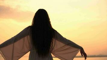 bellissimo ragazza nel un' spiaggia vestito sta su un' tramonto sfondo e si diffonde sua mani nel diverso indicazioni. lento movimento video