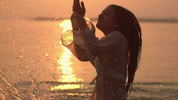 hermosa contento niña con largo oscuro pelo lo hace salpicaduras manos en pie en el río en contra el Dom durante puesta de sol teniendo un bueno humor. lento movimiento. de cerca. hd. contento día video