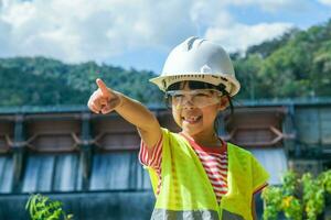 retrato de un pequeño niña ingeniero vistiendo un verde chaleco y blanco casco sonriente felizmente en el antecedentes de el represa. conceptos de ambiental ingeniería, renovable energía y amor de naturaleza. foto