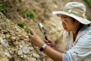 hembra geólogo utilizando aumentador vaso a examinar y analizar roca, suelo, arena en naturaleza. arqueólogos explorar el campo. ambiental y ecología investigación. foto