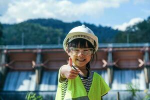Portrait of a little girl engineer wearing a green vest and white helmet smiling happily on the background of the dam. Concepts of environmental engineering, renewable energy and love of nature. photo