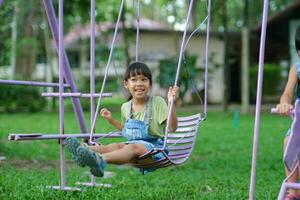 contento asiático niña teniendo divertido jugando en el patio de recreo durante verano. linda pequeño niña balanceo en el patio de recreo con un sonrisa y risa. activo verano ocio para niños. foto