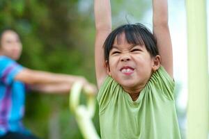 Little girl hanging on the horizontal bar on the playground. Cute young Asian girl hangs bar by hand to exercise at outdoor playground. photo