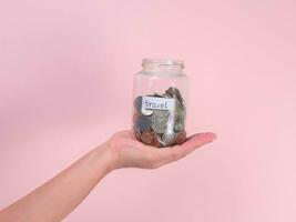 Close-up of young woman's hand holding glass jar with money  inside on pink background. Hand holding a money jar, travel, savings, education, donation. Finance plan concept. photo