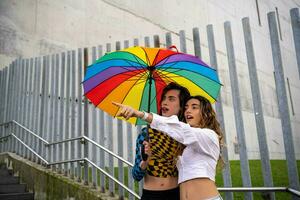 young couple pointing at a point in amazement while holding an umbrella with pride colors. photo