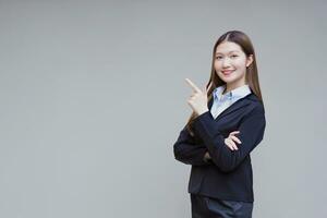 profesional asiático trabajando mujer quien tiene largo pelo usa negro formal traje con azul camisa mientras ella brazo cruce y sonriente presentación en gris antecedentes. foto